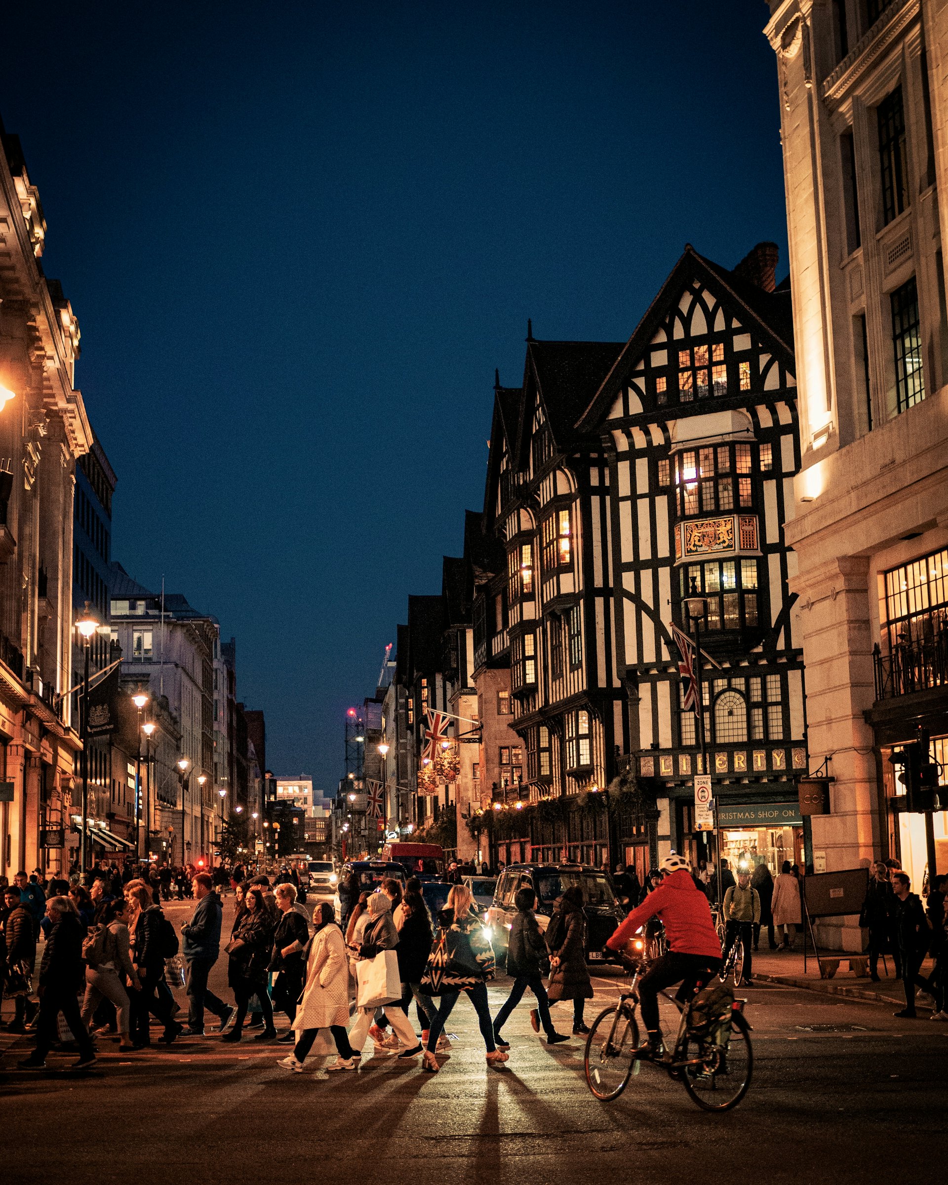 Shoppers on a West End street at dusk beside a Tudor-revival department store